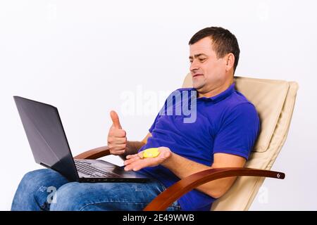 Un homme sourd senior tient une prothèse auditive dans la main et montre le pouce vers le haut à la caméra de l'ordinateur portable tout en étant assis dans une chaise sur un fond blanc. Banque D'Images