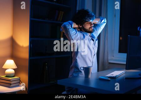 Surtravaillé jeune freelance barbu étirant les mains et commençant à taper sur le clavier la nuit. Banque D'Images