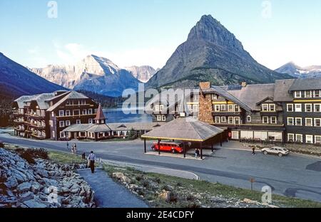 Pendant les pauses d'une journée, un bus rouge vintage vous attend à l'entrée principale de nombreux hôtels Glacier pour les touristes qui effectuent une excursion pittoresque en début de matinée le long de la route montagneuse allant au soleil dans le parc national Glacier, dans le nord-ouest du Montana, aux États-Unis. Le véhicule de 17 passagers fait partie de la flotte de 33 bus touristiques identiques des années 1930 qui sont dotés de toits en toile pour offrir une vue panoramique sur les impressionnants sommets montagneux du parc. Situé sur les rives du lac SwiftCurrent, cet hôtel historique a été construit par le Great Northern Railway en 1914-15 et propose des chambres de style ancien. Banque D'Images