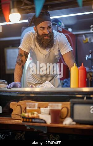jeune beardy hippster travaillant dans le service de restauration rapide, portant un chapeau de cuisinier Banque D'Images
