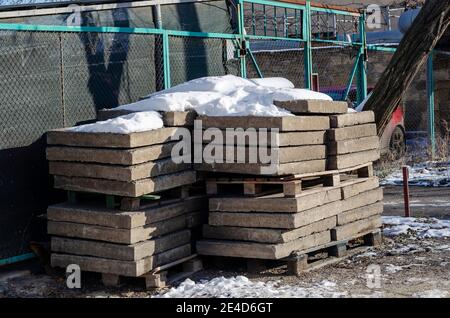 Pile de blocs de béton carrés recouverts de neige. Blocs de béton sur des palettes en bois près de la clôture verte. Jour d'hiver ensoleillé. Personne. Banque D'Images