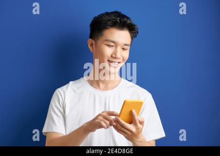 Étudiant vêtu décontracté, isolé sur fond bleu, regardant l'écran du smartphone, parcourant des pages Web et souriant tout en discutant avec ven Banque D'Images
