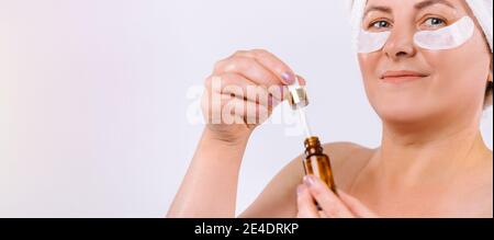 Bannière,format long. Une photo rognée d'une femme plus âgée tient un sérum de levage avec une pipette devant elle. Arrière-plan blanc et espace publicitaire latéral vide. Banque D'Images