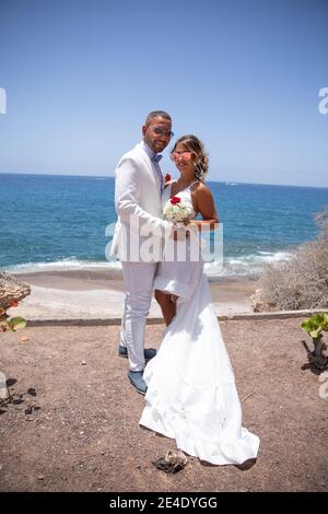 Couple récemment marié tenant le bouquet de mariage, ils sont élégamment habillés en blanc avec des lunettes de soleil et ont la mer derrière eux. Pho pleine longueur Banque D'Images