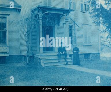 Archive américaine portrait cyanotype d'une maison de galets à Port Byron avec un vieil homme avec une barbe assis sur les marches du porche et sa femme debout à côté de lui. Prenez la fin du XIXe siècle à Port Byron, NY, États-Unis Banque D'Images