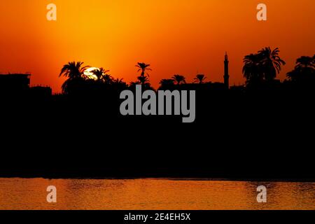 Coucher de soleil sur la rive du Nil, Louxor, Égypte avec silhouettes d'une mosquée et palmiers sur la ligne d'horizon Banque D'Images