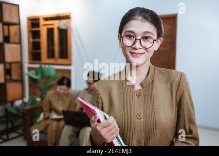 enseignante en uniforme de fonctionnaire portant des lunettes souriant pendant portant un livre avec l'arrière-plan d'une équipe de enseignants travaillant à domicile Banque D'Images