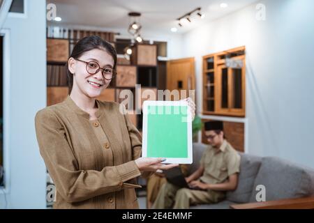 enseignante dans un uniforme de fonctionnaire portant des lunettes souriantes tout en montrant une tablette numérique avec un arrière-plan de travail de chez vous Banque D'Images