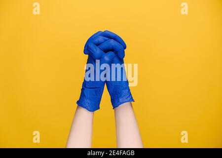 Gros plan une femme méconnaissable en gants médicaux bleus tient les mains dans la prière à dieu, méditant, priant pour la santé, isolée sur studio jaune Banque D'Images