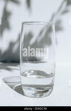 Verre d'eau debout devant un mur blanc avec des ombres de feuilles sur un jour lumineux et ensoleillé Banque D'Images