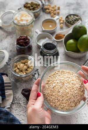 Bol en verre rempli de céréales d'avoine tenu par les mains de femme entouré d'ingrédients de granola dans des bols. Banque D'Images