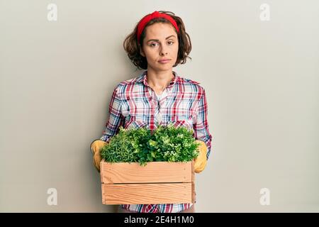 Jeune femme brune portant des vêtements de jardinier tenant pot de plante en bois détendu avec une expression sérieuse sur le visage. Simple et naturel regarder le venu Banque D'Images