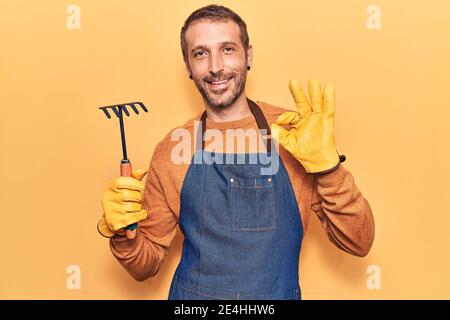 Jeune homme beau portant un tablier de jardinier et des gants tenant le râteau faisant signe ok avec les doigts, souriant sympathique gestante excellent symbole Banque D'Images