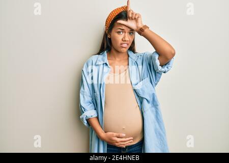 Belle femme hispanique s'attendant à un bébé, touchant le ventre enceinte rendant amusant des gens avec les doigts sur le front faisant le geste perdant mocking et inssul Banque D'Images
