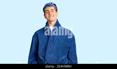 Jeune homme beau portant un masque de sommeil et un peignoir d'aspect positif et se tenir debout et sourire avec un sourire confiant dents Banque D'Images
