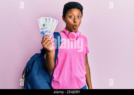 Jeune fille afro-américaine portant un sac à dos étudiant tenant des billets colombiens pesos effrayés et étonnés par la bouche ouverte pour la surprise, incrédulité visage Banque D'Images