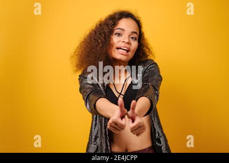 Mains avant et pouces vers le haut bonne fille afro-américaine avec belle coiffure posant le sourire regardant à l'extérieur avec la main sur les hanches portent du noir brillant Banque D'Images