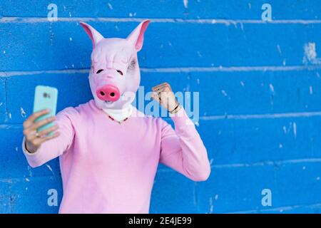 Jeune homme dans le masque de porc montrant le poing en prenant le selfie contre le mur bleu Banque D'Images
