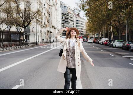 Femme tendance avec les yeux fermés tenant le chapeau tout en étant debout rue Banque D'Images