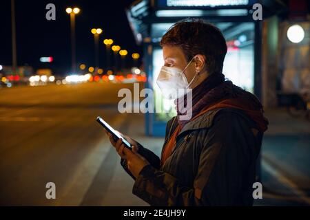 Femme portant un masque FFP2 utilisant un smartphone en attendant bus en ville Banque D'Images