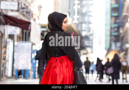 Portrait de la jeune belle femme portant le hijab noir debout sur le trottoir et en regardant l'appareil photo par-dessus l'épaule Banque D'Images