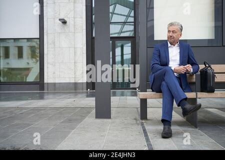Homme d'affaires senior attendant assis avec les jambes croisées dans le bus arrêt en ville Banque D'Images