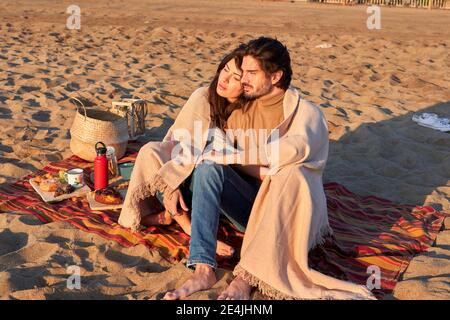 Jeune couple dans une couverture regardant se reposer tout en étant assis sur la plage Banque D'Images