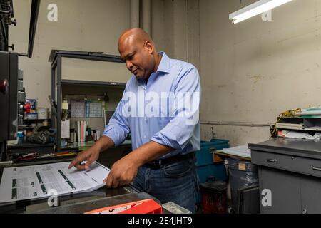 Un superviseur souriant qui regarde loin tout en tenant la planche à papier près d'une machine à imprimer dans l'atelier Banque D'Images