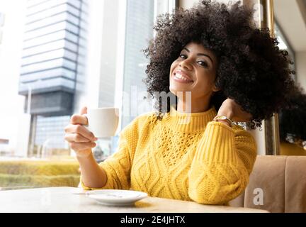 Jeune femme avec la main dans les cheveux tenant la tasse de café pendant assis au café Banque D'Images