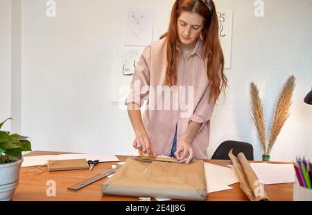 Une artiste féminine applique du ruban adhésif sur du papier brun pendant la préparation de l'emballage dans la salle de séjour Banque D'Images