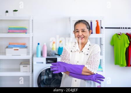 Femme asiatique aînée portant des gants en caoutchouc et des bras croisés dans la salle de blanchisserie avec fierté, regardant la caméra, concept de nettoyage de printemps Banque D'Images