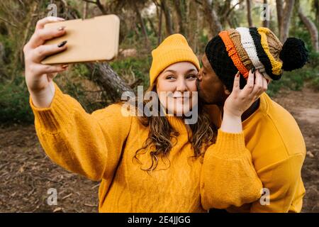 Homme embrassant femme prenant selfie par téléphone portable en étant debout en forêt Banque D'Images