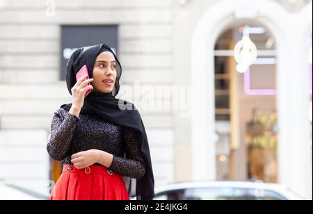 Portrait d'une jeune belle femme portant un hijab noir parlant sur un smartphone au milieu de la rue Banque D'Images