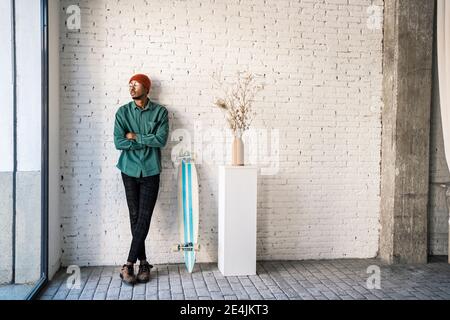 Homme réfléchi aux bras croisés debout par planche à roulettes contre le blanc mur de briques Banque D'Images