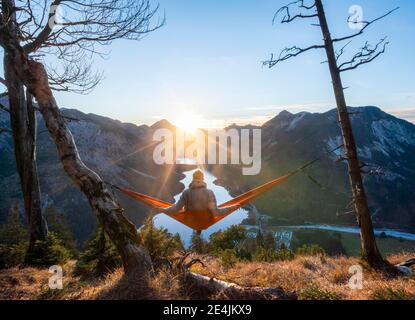 Jeune homme assis dans un hamac rouge, vue panoramique sur les montagnes avec lac, étoile de soleil, coucher de soleil, Plansee, Tyrol, Autriche Banque D'Images