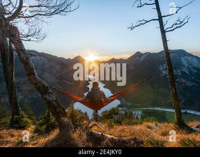 Jeune homme assis dans un hamac rouge, vue panoramique sur les montagnes avec lac, étoile de soleil, coucher de soleil, Plansee, Tyrol, Autriche Banque D'Images