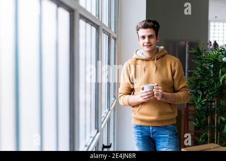 Jeune homme souriant tenant une tasse de café debout près de la fenêtre dans une nouvelle maison Banque D'Images