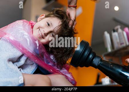 Fille souriante appréciant tout en main de femme coiffeur utilisant le sèche-linge sur les cheveux mouillés du client dans le salon Banque D'Images