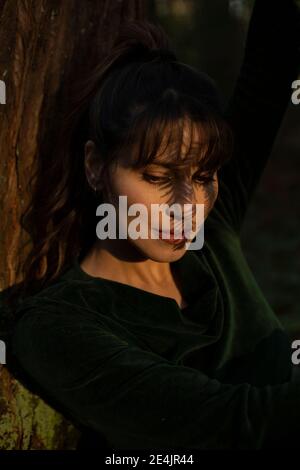 Femme regardant vers le bas tandis que l'ombre tombe sur le visage tout en se penchant sur le tronc de l'arbre en plein soleil Banque D'Images