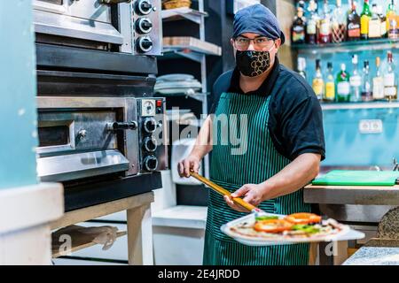 Portrait d'un chef masculin portant un masque de protection tenant le pelage avec une pizza fraîchement préparée Banque D'Images