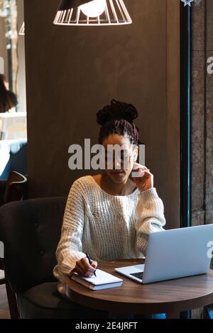Belle jeune femme écrivant dans un journal tout en étant assise avec un ordinateur portable au café Banque D'Images