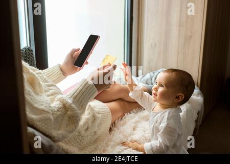 Mignonne fille de bébé atteignant à la carte de crédit étant tenue par mère assise près de la fenêtre à la maison Banque D'Images