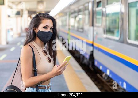 Jeune femme portant un masque facial avec un téléphone portable debout sur la plate-forme Banque D'Images