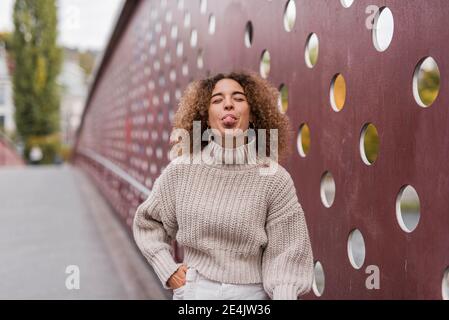 Jeune femme afro ludique qui colle la langue tout en se tenant contre un mur métallique Banque D'Images