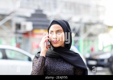 Portrait d'une jeune belle femme portant un hijab noir parlant sur un smartphone au milieu de la rue Banque D'Images