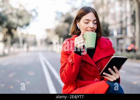 Jeune femme souriante en veste d'hiver ayant le café tout en utilisant tablette numérique sur la route Banque D'Images