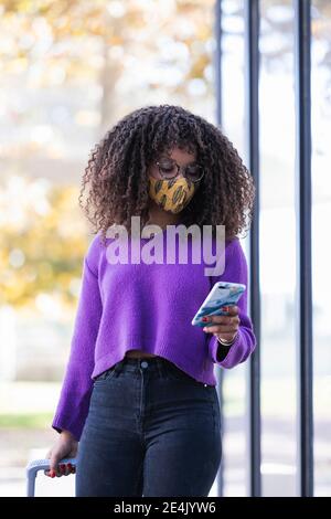 Jeune femme à la mode utilisant un téléphone portable tout en marchant avec des bagages Pendant COVID-19 Banque D'Images