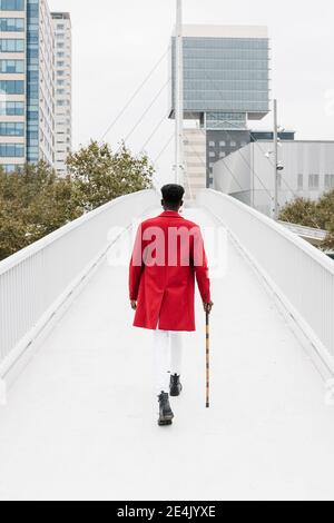 Homme portant une veste rouge marchant avec une canne sur le pont en ville Banque D'Images