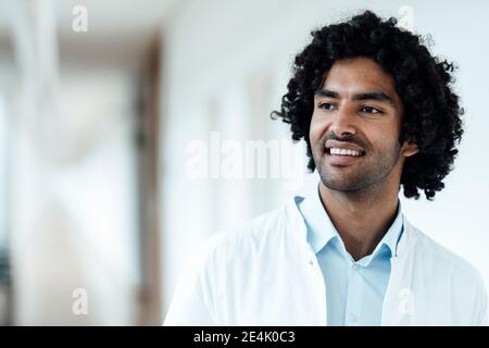Homme souriant professionnel de la santé avec des cheveux bouclés noirs regardant loin à l'hôpital Banque D'Images