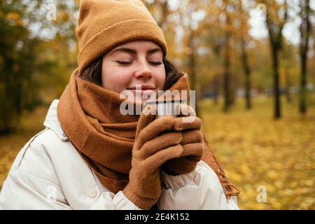 Jeune femme souriante avec les yeux fermés odeur de thé en automne stationnement Banque D'Images